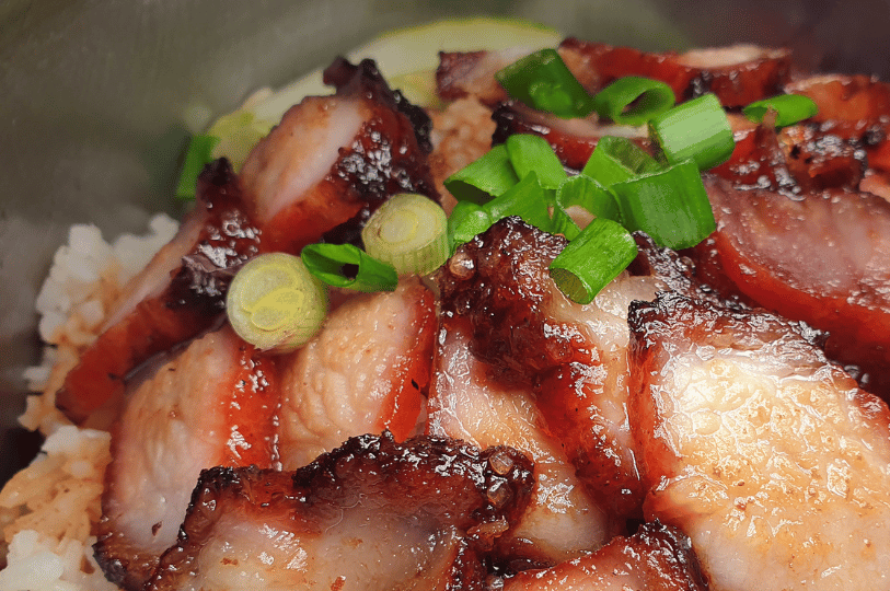 Art-of-the-Tease- (3) A decadent close-up of sliced Char Siu (BBQ pork) resting on a bed of white rice. The pork features a sticky, caramelized red glaze and charred, crispy edges, garnished with bright green scallion rings.
