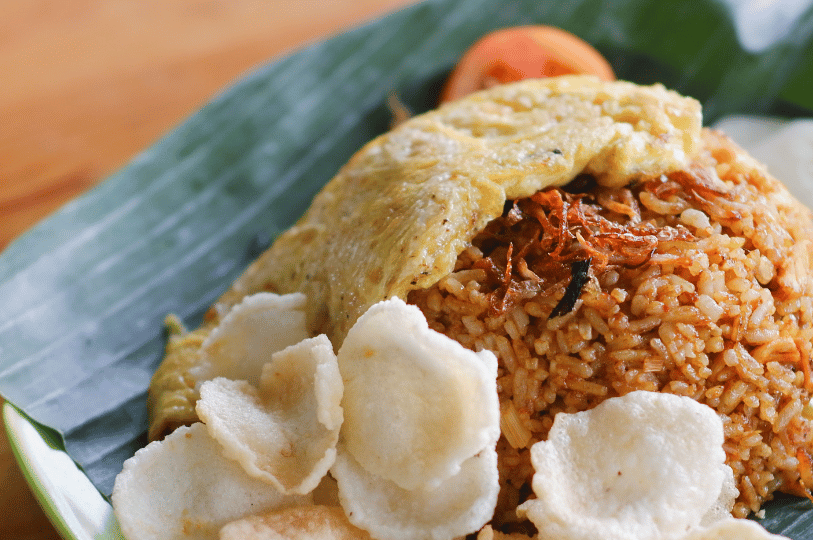 Art-of-the-Tease- (5) A vibrant plate of fried rice presented on a traditional banana leaf. The mound of seasoned rice is topped with crispy fried shallots, a golden fried egg, and a side of white shrimp crackers.