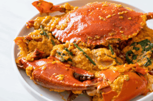 A high-angle shot of a large red chili crab in a white bowl, coated in a thick, eggy tomato gravy. The bright orange-red shell contrasts with the yellow sauce and fresh green herbs, demonstrating high-key lighting that emphasizes the dish's volume.