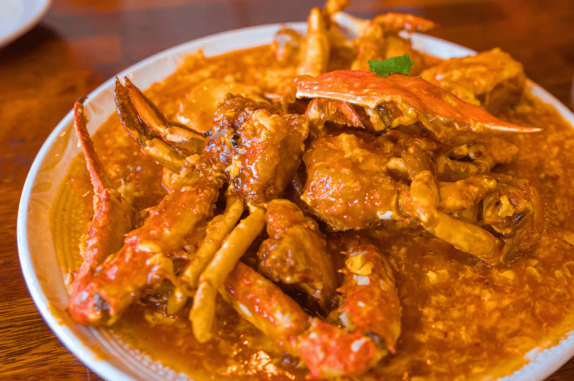 A warm-toned, eye-level shot of chili crab served on a white plate. The lighting highlights the glistening, semi-translucent quality of the savory sauce and the intricate shapes of the crab legs, set against a blurred wooden background.