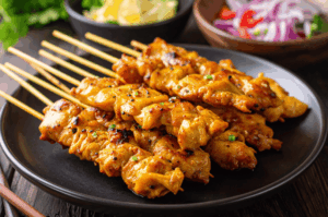 A close-up, low-angle shot of a plate of charred Chicken Satay skewers. The skewers are arranged diagonally to create leading lines, with a blurred background of red onions and cucumbers emphasizing the texture of the peanut-glazed meat.