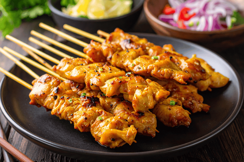 Composition- (1) A close-up, low-angle shot of a plate of charred Chicken Satay skewers. The skewers are arranged diagonally to create leading lines, with a blurred background of red onions and cucumbers emphasizing the texture of the peanut-glazed meat.
