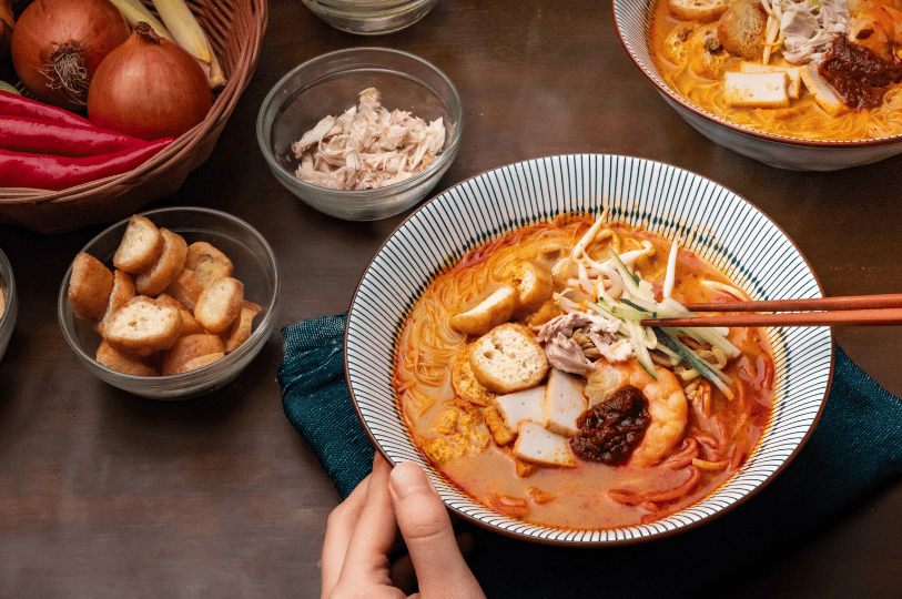 Composition- (2) A top-down "flat lay" perspective of a bowl of Singapore Laksa using rule of thirds. A hand enters the frame holding a pair of chopsticks, adding a human element. Surrounding ingredients like lemongrass, shallots, and tofu puffs are arranged to frame the central bowl.