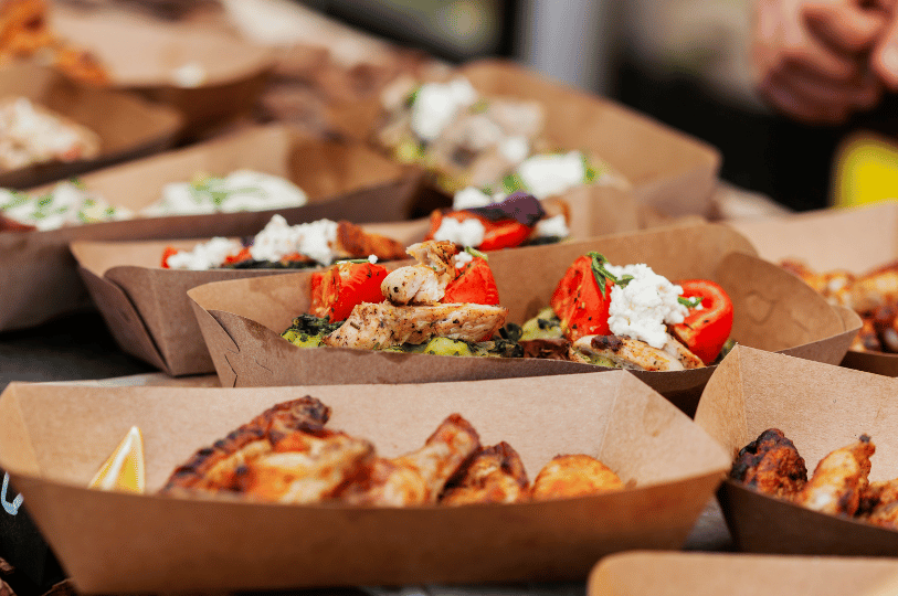 Composition- (5) A mid-range shot of multiple cardboard food trays containing grilled chicken and tomatoes making use of layering and depth. The trays are organized in rows, using a rhythmic pattern and warm lighting to showcase street-food-style catering.