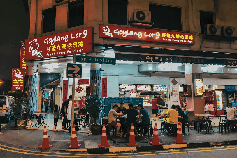 Geylang- (1) Nighttime street scene outside Geylang Lor 9 Fresh Frog Porridge, with diners at outdoor tables under glowing red signage, capturing the district’s lively, late-night allure and culinary boldness.