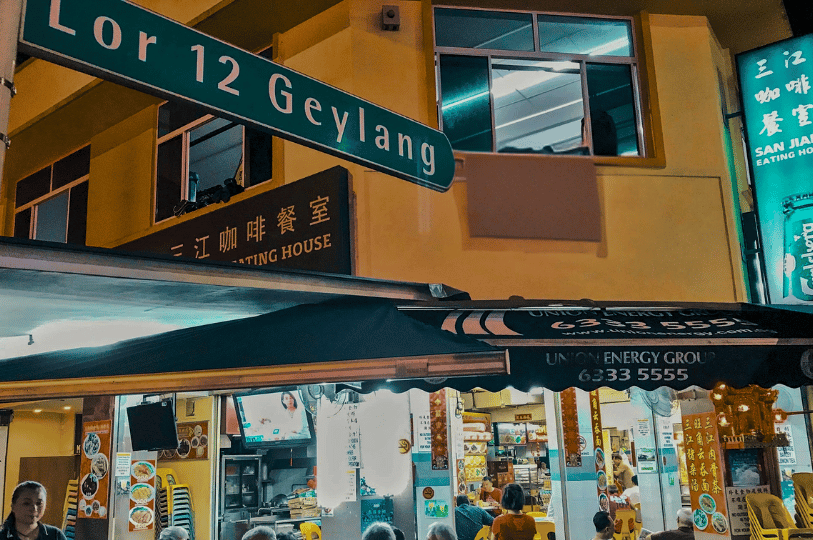 Geylang- (2) Bustling nighttime view of San Jiang Eating House on Lorong 12 Geylang, with diners gathered under bright signage and food displays, evoking the district’s irresistible mix of flavor, energy, and street-side charm.