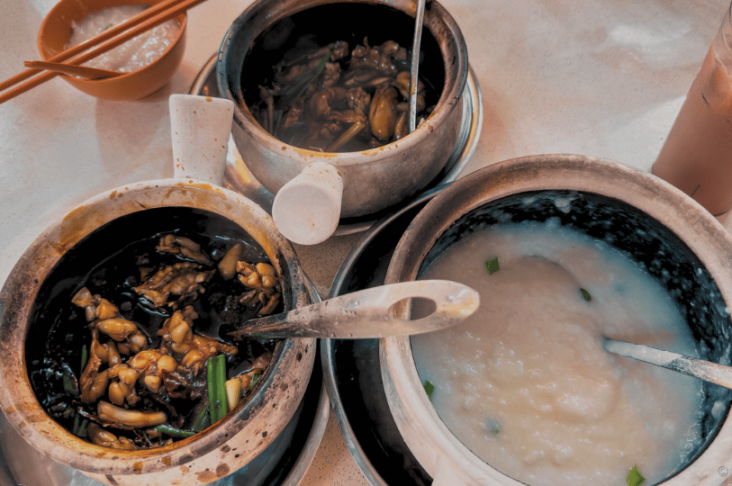 Geylang- (3) Three clay pots of frog leg dishes and porridge on a table, showcasing Geylang’s signature delicacy—rich, savory frog legs in dark sauce beside creamy congee—an emblem of the district’s daring and delicious food culture.
