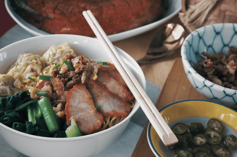 Geylang- (4) Close-up of a noodle bowl topped with roasted pork, dumplings, and greens, surrounded by chili soy, minced condiments, and charred meat—an indulgent spread that captures Geylang’s unapologetic devotion to bold, late-night flavors.