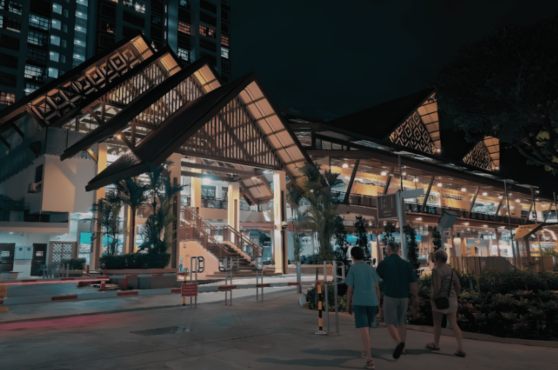 Geylang- (6) Illuminated night view of Geylang Serai Market’s striking pitched-roof architecture, as visitors approach its glass-fronted entrance—blending Malay heritage with modern design in the heart of Singapore’s cultural foodscape.