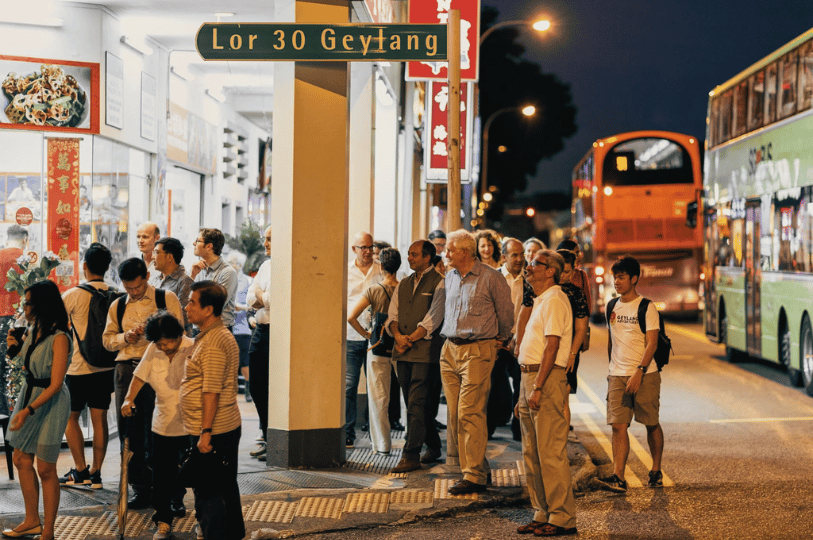 A group of people gathered under a green street sign for Lor 30 Geylang in Singapore at night, with a classic double-decker bus passing by in the background.