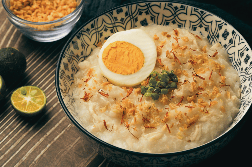 A bowl of savory Cantonese-style chicken porridge garnished with a hard-boiled egg, fried shallots, chopped spring onions, and ginger strips, served with fresh lime.