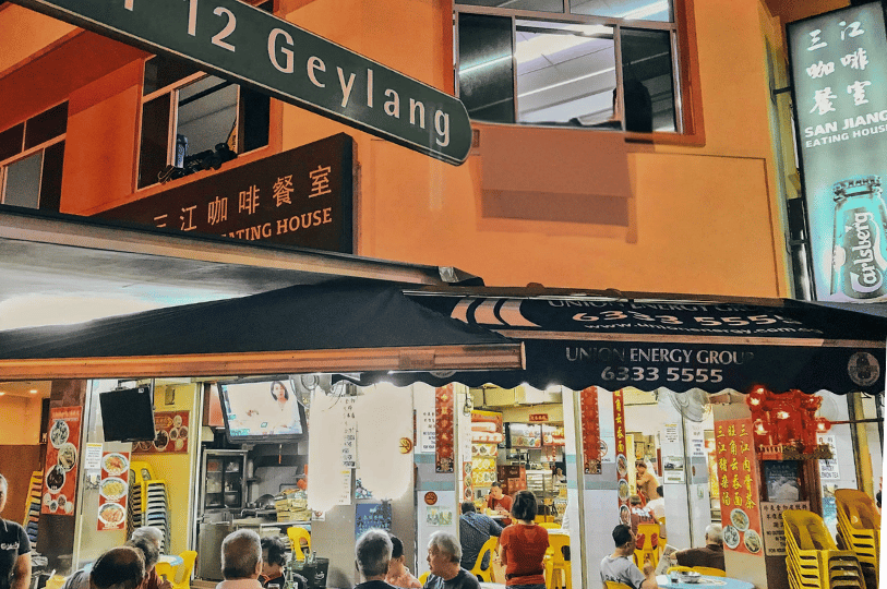 The vibrant exterior of San Jiang Eating House at Lor 12 Geylang, showing an open-air coffee shop (kopitiam) with patrons dining under a yellow-lit awning.