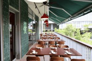 An elevated, open-air balcony dining area of an Asian restaurant in Holland Village, showcasing wooden tables neatly set with traditional bowls and spoons, red pendant lights, and a green tiled wall accented by a glowing green neon Chinese character.