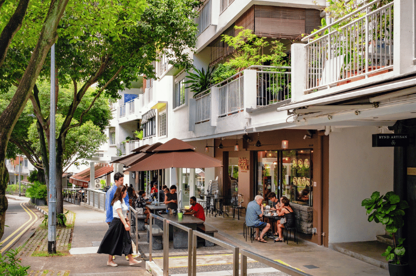 Holland-Village- (2) A charming tree-lined street in Holland Village where a modern "Baker & Cook" cafe sits below traditional white apartments, with patrons enjoying alfresco dining.