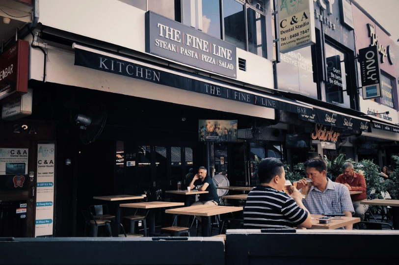 Street-level view of patrons relaxing with drinks at the outdoor seating area of The Fine Line, a Western-style bar and restaurant featuring dark awnings, street-side wooden tables, and a relaxed pub atmosphere in Holland Village.