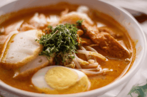 A close-up shot of a creamy bowl of Laksa topped with a pile of finely shredded laksa leaves. The rich orange broth surrounds half a hard-boiled egg, thick fish cake slices, bean sprouts, and fried tofu puffs.