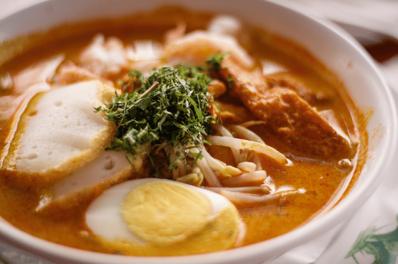 A close-up shot of a creamy bowl of Laksa topped with a pile of finely shredded laksa leaves. The rich orange broth surrounds half a hard-boiled egg, thick fish cake slices, bean sprouts, and fried tofu puffs.