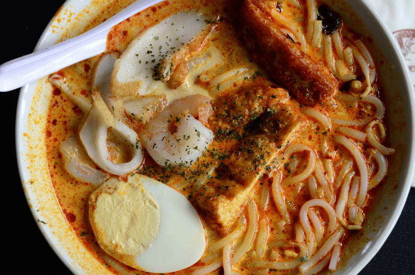 An overhead view of a bowl of curry laksa featuring thick rice noodles submerged in a vibrant, coconut-based orange broth. The dish is packed with shrimp, fish cake, a hard-boiled egg, and fried tofu, all garnished with dried herbs.