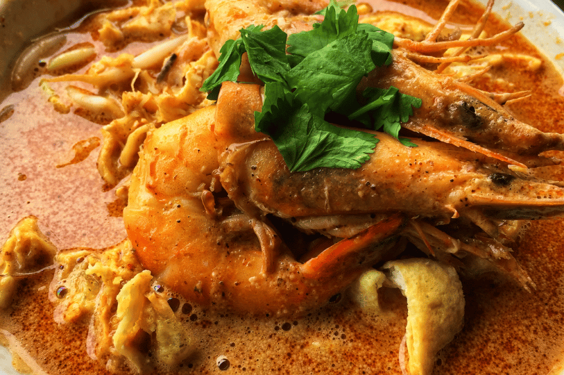 A macro shot focusing on several large, succulent prawns resting atop a thick, bubbly laksa broth. A fresh sprig of cilantro sits on the prawns, highlighting the contrast between the seafood and the spicy, textured gravy.