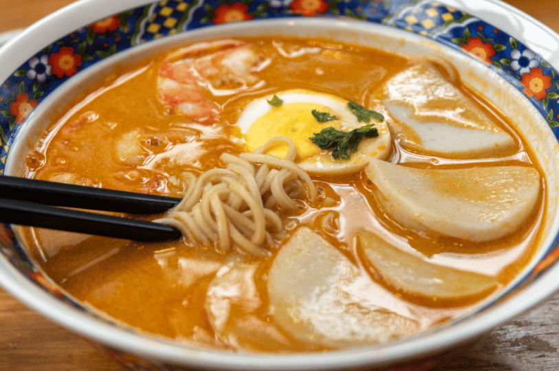 A bowl of laksa served in a decorative blue and white floral porcelain bowl. A pair of black chopsticks lifts a bundle of thin noodles from the creamy broth, which is garnished with fish cakes, shrimp, and a hard-boiled egg.