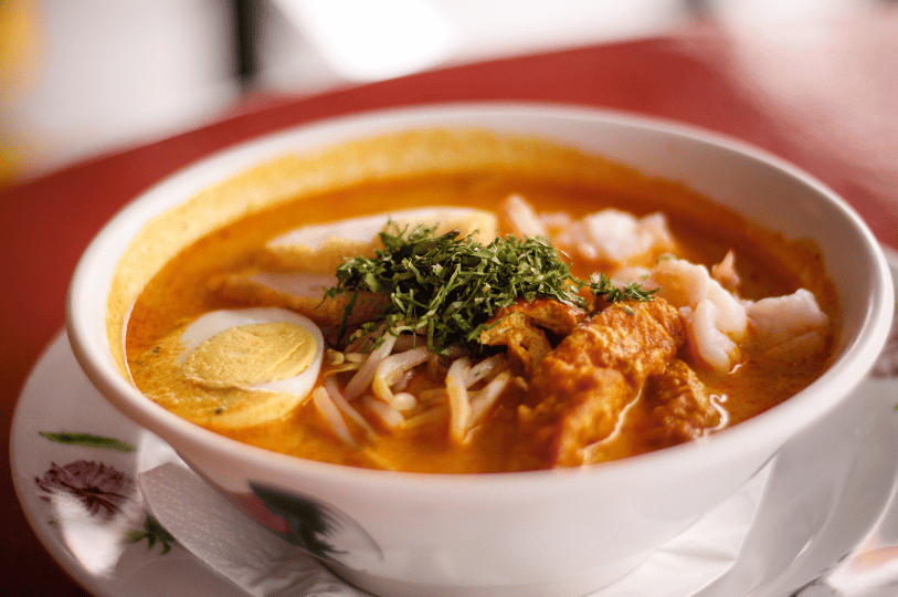 A side-angle view of a steaming bowl of laksa, emphasizing the depth of the creamy coconut curry broth. The bowl is topped with bean sprouts, fried tofu, and a generous mound of chopped laksa leaves, served on a saucer.
