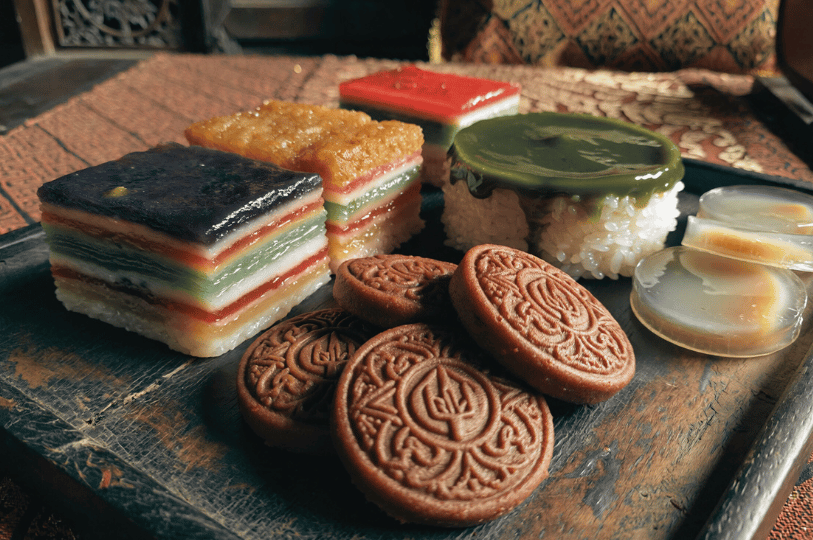 A high-angle, close-up shot of various Peranakan kueh (sweets) on a rustic wooden tray. The selection includes a multi-colored Kueh Lapis Sagu, a glutinous rice cake topped with green pandan custard (Kueh Salat), and embossed brown cookies. The lighting is warm and directional, highlighting the intricate textures and vibrant layers against a dark, moody background.