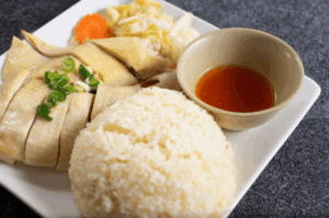 A close-up, high-angle shot of Hainanese Chicken Rice served on a white plate. It features a neat mound of fragrant rice, sliced poached chicken garnished with scallions, and a small bowl of bright chili sauce.