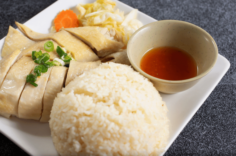 Tiong-Bahru- (1) A close-up, high-angle shot of Hainanese Chicken Rice served on a white plate. It features a neat mound of fragrant rice, sliced poached chicken garnished with scallions, and a small bowl of bright chili sauce.