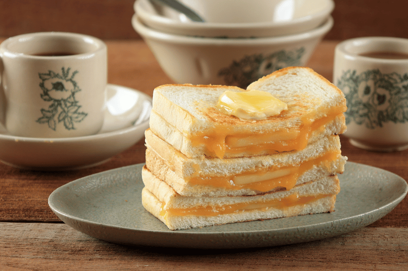 Tiong-Bahru- (3) A warm, inviting shot of Kaya Toast stacked on a blue plate. Thick slabs of butter and golden kaya jam ooze from between toasted white bread, with traditional floral-patterned coffee cups in the blurred background.
