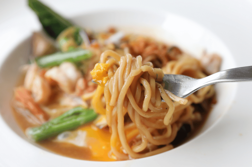 Tiong-Bahru- (5) A dynamic "action shot" of a fork lifting a swirl of saucy noodles from a white bowl. The focus is on the glossy texture of the noodles and a soft-boiled egg yolk mixing into the savory broth.