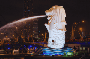 The iconic Merlion statue in Singapore illuminated at night, spraying a fountain of water into the bay against a backdrop of glowing city skyscrapers and streetlights.