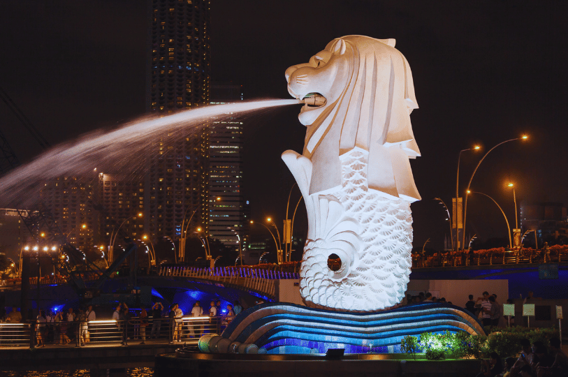 The iconic Merlion statue in Singapore illuminated at night, spraying a fountain of water into the bay against a backdrop of glowing city skyscrapers and streetlights.