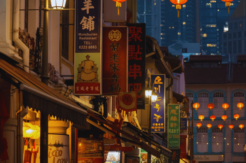 A vertical shot of a narrow Singaporean street at dusk, filled with glowing traditional red lanterns and layered vertical shop signs for various Chinese restaurants and bars.