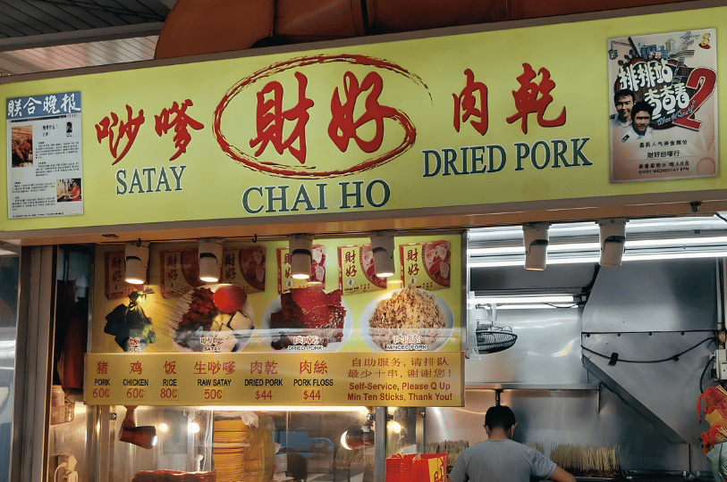 A brightly lit hawker stall named "Chai Ho Satay & Dried Pork" featuring a yellow sign with red Chinese calligraphy. The menu displays prices for pork, chicken, and rice, along with images of grilled satay and bak kwa (dried pork).