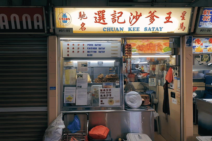 The storefront of "Chuan Kee Satay," a traditional Singaporean hawker stall. A simple white signboard with red and blue lettering lists pork, chicken, and mutton satay. Bundles of raw skewers are visible through the glass display.