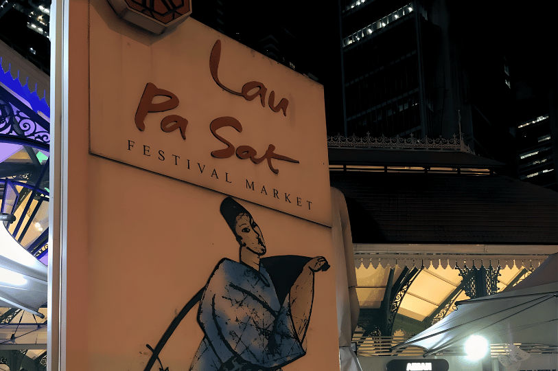 A low-angle shot of the iconic "Lau Pa Sat Festival Market" sign at night. The warm glow of the historic Victorian-style cast-iron structure is visible in the background against the dark city skyline.