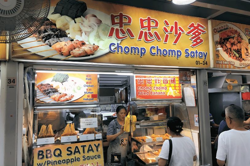 A vibrant stall named "Chomp Chomp Satay" with a large orange and yellow banner. The display highlights "BBQ Satay with Pineapple Sauce" and shows a vendor preparing orders behind a glass counter filled with skewers.