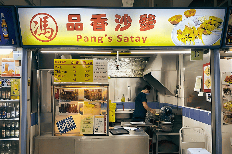 The "Pang’s Satay" hawker stall featuring a clean, backlit yellow sign. A price list shows pork, chicken, and mutton skewers for 60 cents each. A vendor in a face mask is seen working in the stainless-steel kitchen area.