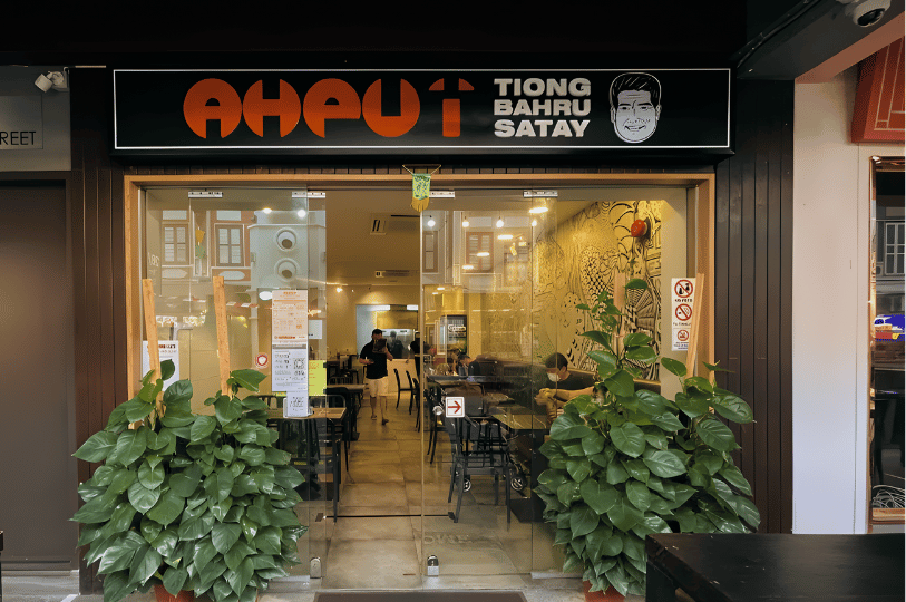 The modern glass storefront of "Ah Pui Tiong Bahru Satay." The black signage features a minimalist orange logo and a caricature of the famous founder, overlooking an indoor dining area with warm lighting.