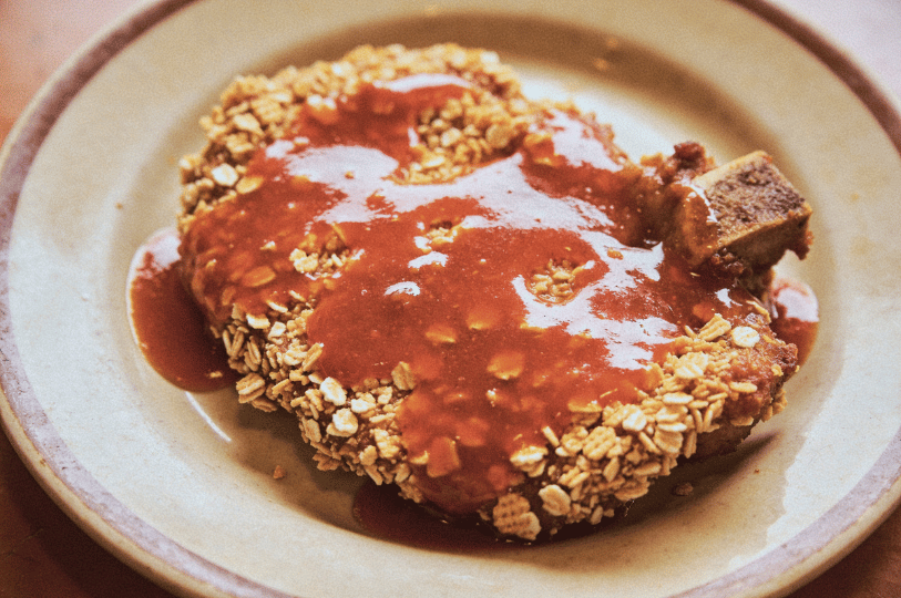 Colonial-Photoshoot- (3) A top-down food photography shot of a breaded cutlet— a Hainanese pork chop—heavily coated in crushed oats or crackers and smothered in a vibrant tomato-based gravy. The dish is served on a classic cream-colored ceramic plate with a simple brown rim.