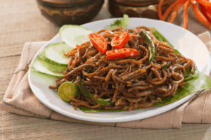 A vibrant plate of Hokkien Mee served on a banana leaf. The dark, saucy noodles are garnished with sliced red chilies, green chilies, a half-cut calamansi lime, and fresh cucumber slices, showcasing a classic Southeast Asian street food staple.