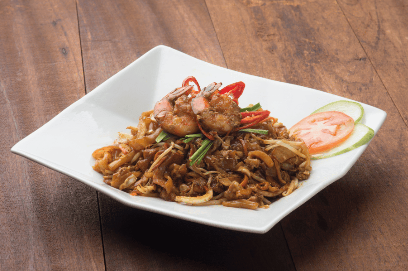 A high-angle shot of Char Kway Teow served in a modern white square bowl on a rustic wooden table. The stir-fried flat rice noodles are topped with large succulent prawns, bean sprouts, and chives, reflecting a fusion of traditional ingredients and contemporary plating.