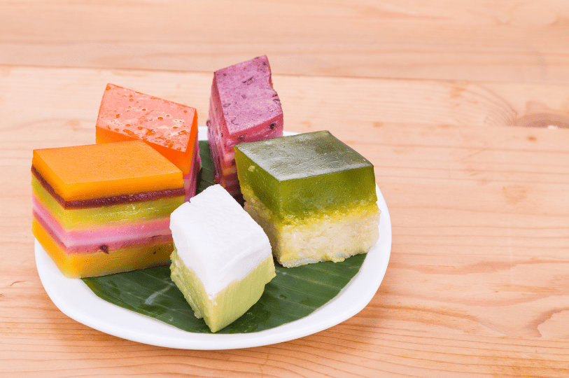 A colorful assortment of Kueh (traditional Southeast Asian steamed cakes) arranged on a white plate with a banana leaf liner. The display includes layered rainbow cakes, a green and white coconut milk cake (Kueh Talam), and a purple-speckled variety, highlighting the geometric beauty of traditional desserts.