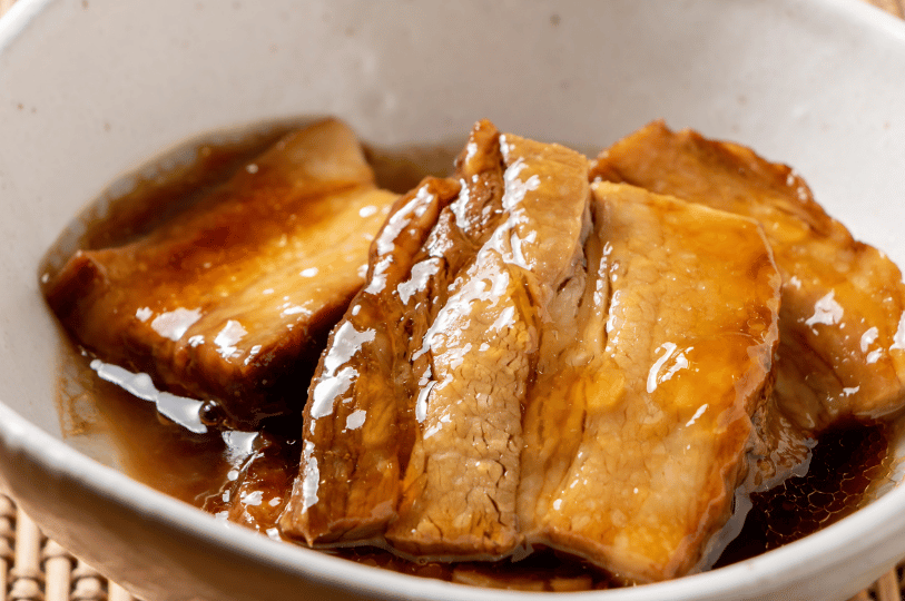 A detailed shot of Braised Pork Belly in a ceramic bowl. The thick, glistening slices of meat are coated in a dark soy sauce glaze, showing the tender fat layers and rich marination.