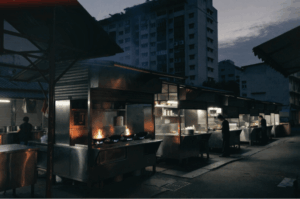 A wide shot of a row of food stalls at a morning market before sunrise. Bright orange flames flare up from woks in the foreground, contrasting with the cool, dark blue hues of the early morning sky and a residential apartment building in the background.