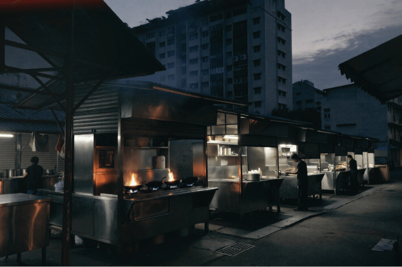 A wide shot of a row of food stalls at a morning market before sunrise. Bright orange flames flare up from woks in the foreground, contrasting with the cool, dark blue hues of the early morning sky and a residential apartment building in the background.