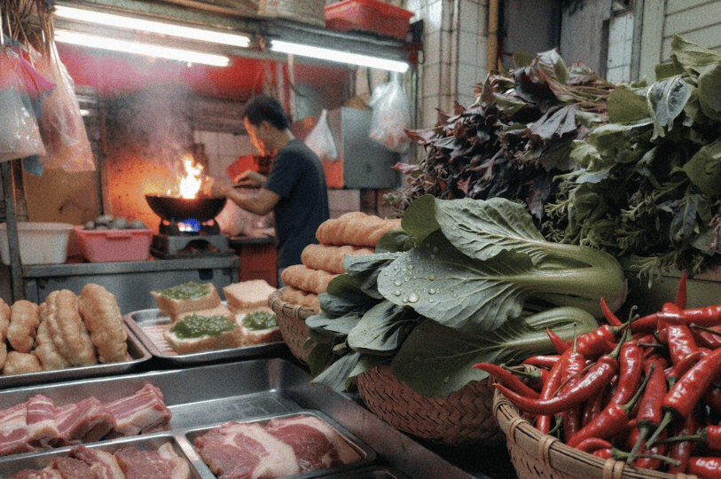 Dawn-Morning-Market-Food-Blog- (2) A vibrant close-up of a market stall featuring baskets of fresh red chilies and green leafy bok choy in the foreground. In the soft-focus background, a vendor is busy cooking over a fiery wok next to trays of sliced pork and toast topped with green kaya jam.