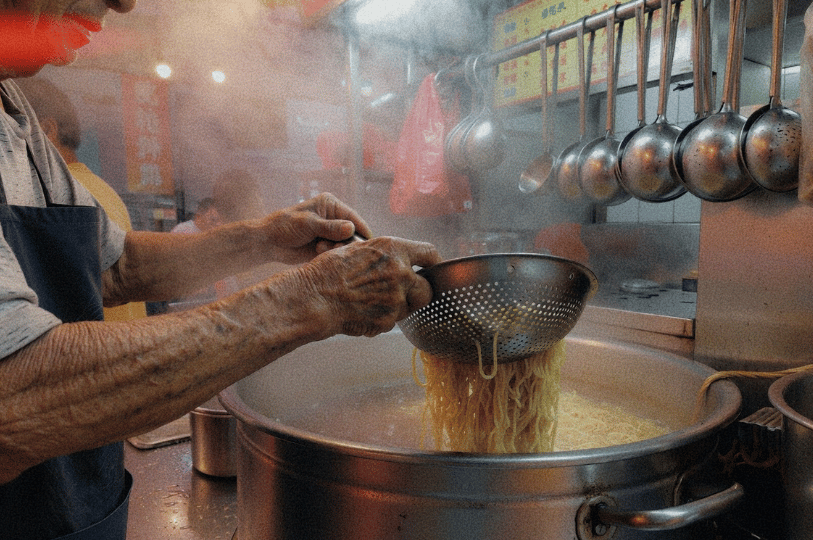 Dawn-Morning-Market-Food-Blog- (3) A close-up, atmospheric shot of a street food vendor’s weathered hands using a metal strainer to lift a large portion of steaming yellow noodles from a massive boiling pot. Soft morning light and steam fill the frame, highlighting the texture of the noodles.