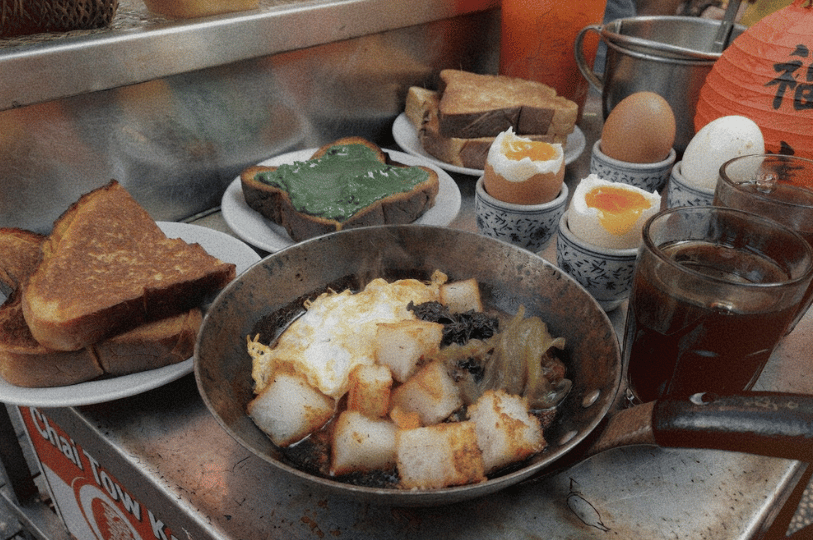 Dawn-Morning-Market-Food-Blog- (4) A traditional Nanyang breakfast spread on a stainless steel table. The meal includes a small pan of fried carrot cake (chai tow kway) with egg, several slices of thick-cut kaya toast, soft-boiled eggs in ceramic cups, and two glasses of dark kopi (coffee).