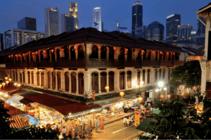 An elevated evening view of a historic multi-story shophouse in Chinatown, adorned with glowing red lanterns, with bustling street market stalls in the foreground and the modern Singapore skyline rising in the background.